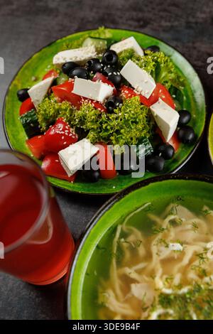 Salate mit Salat, Tomaten, Gurken, Oliven und Käse werden auf Tellern serviert. Eine Schüssel Suppe und ein Glas rotes Getränk stehen in der Nähe, was hervorhebt Stockfoto