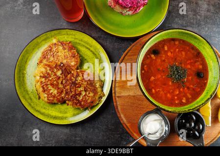 An einem Tisch werden herzhafte ukrainische Küche mit knusprigen Kartoffelpfannkuchen auf einem Teller, rotem Borschtsch in einer Schüssel und Toppings wie Sauerrahm und Oliven in der Nähe präsentiert Stockfoto