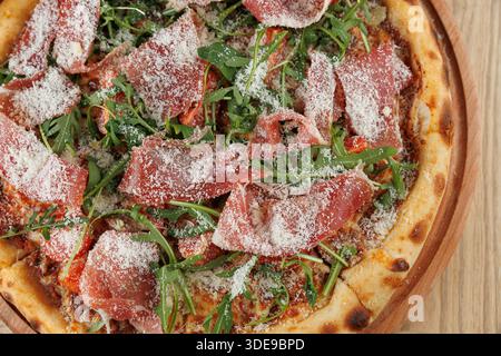 Eine frische Pizza mit Schinkenscheiben, Rucola und geriebenem Käse wird auf ein Holzbrett gelegt. Die Kruste ist goldfarben und knusprig, bereit zum Servieren Stockfoto