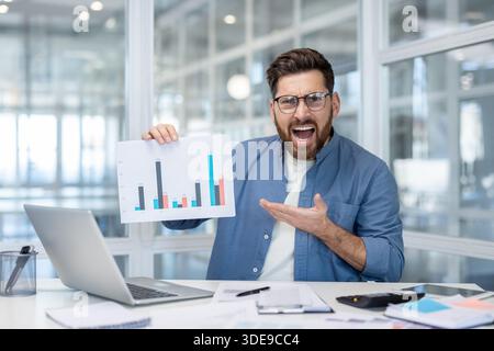 Ein gestresster Geschäftsmann sitzt an einem Schreibtisch, hält ein Balkendiagramm mit negativer Leistung, blickt in die Kamera und brüllt frustriert durch finanzielle Verluste und schlechte Ergebnisse Stockfoto