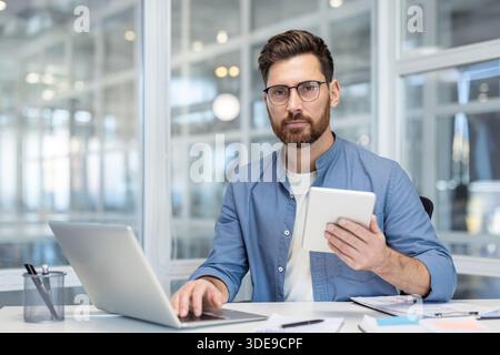 Professioneller Mann mit Brille und Bart, der in einem hellen, modernen Büro am Schreibtisch arbeitet, ein Notebook verwendet und ein Tablet hält, während er Daten und Planungsstrategien analysiert und dabei selbstbewusst und konzentriert wirkt Stockfoto