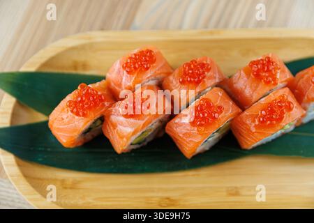 Frisch zubereitete Sushi-Brötchen werden auf einer Holzplatte serviert. Jedes Brötchen enthält Lachsscheiben und wird mit hellrotem Fischrogen belegt. Grüne Blätter Stockfoto