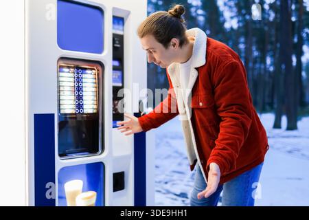 Überraschter junger Mann, der im Winter in einem verschneiten Park einen Kaffeeautomaten ansah. Hochwertiges Foto || Modell veröffentlicht Stockfoto