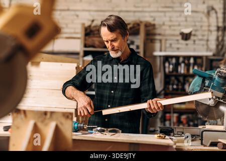 Zimmermann hält eine Holzdiele und schaut auf Maßband in einer Werkstatt || Model veröffentlicht Stockfoto