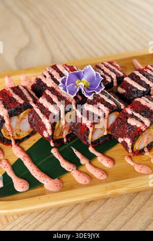 Sushi-Brötchen sind wunderschön auf einer Bambusplatte angeordnet. Die Brötchen haben leuchtend rote Beläge und einen Hauch Soße, gekrönt mit einer violetten Blume f Stockfoto