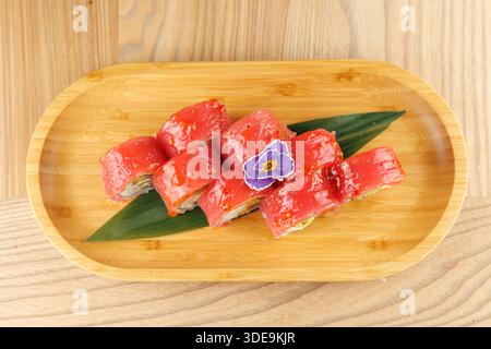 Auf einem Holzteller stehen lebendige Sushi-Brötchen, garniert mit einer violetten Blume. Das Sushi enthält frische Zutaten, die japanische Küche zum Ausdruck bringen Stockfoto