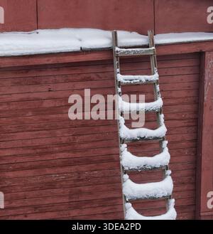 Eine alte Holzleiter, die mit Schnee bedeckt ist, lehnt sich an eine rote Scheunenwand mit einer schneebedeckten Dachkante. Stockfoto