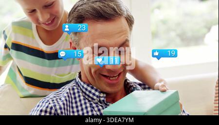 Mann, der zu Hause lächelt, kariertes Hemd trägt, türkisfarbenes Geschenk mit sozialen Ikonen hält Stockfoto