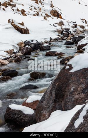 Im Winter fließt ein kalter Gebirgsbach zwischen schneebedeckten Felsen und Felsbrocken. Natürliche Alpenlandschaft mit klarem Wasser, Eis und dramatischer saisonaler A Stockfoto