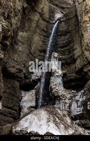 Kleiner Wasserfall teilweise gefroren in einer schmalen Felsschlucht. Winterberglandschaft mit Eisformationen und fließendem Wasser. Stockfoto