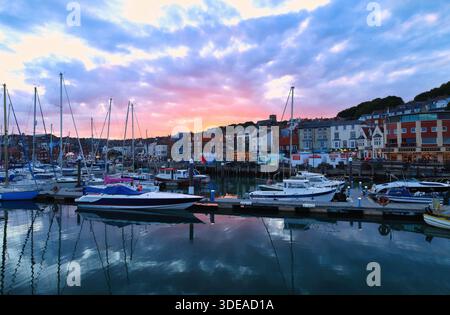 Yachten legen bei Sonnenuntergang im Hafen von Scarborough an, wobei die Küste der Stadt eine malerische Kulisse unter dem dramatischen Abendhimmel bildet – Yorkshire, Großbritannien Stockfoto