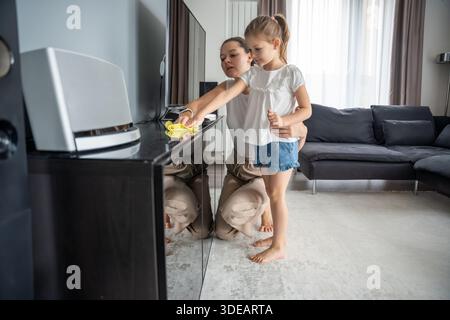Mutter und Tochter, die den Fernseher abwischen, stehen zu Hause zusammen. Haushaltsalltag und Mitverantwortung im realen Innenraum Stockfoto