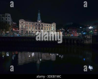 Bilbao Rathausfassade beleuchtet bei Nacht, reflektiert auf dem ruhigen Wasser des nervion Stockfoto