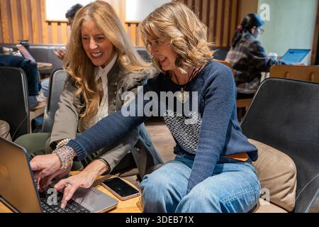 Zwei Freunde sitzen eng zusammen in einem einladenden Café, teilen Ideen und lachen, während sie an einem Laptop arbeiten. Ihre fröhliche Interaktion schafft ein vib Stockfoto