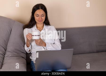 Lächelnde Frau, die mit Laptop sitzt und eine weiße Tasse hält. Stockfoto