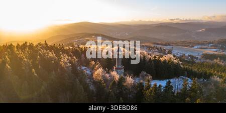 Der Stepanka-Aussichtsturm steht hoch im Isergebirge, wenn die Sonne untergeht, und wirft goldenes Licht über verschneite Bäume und die Landschaft darunter. Besucher genießen die Winterlandschaft. Stockfoto