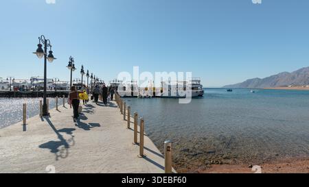 Dahab, Ägypten - 5. Februar 2024: Touristen gehen entlang des Piers zu weißen Vergnügungsyachten und Booten, die im Roten Meer vertäut sind Stockfoto