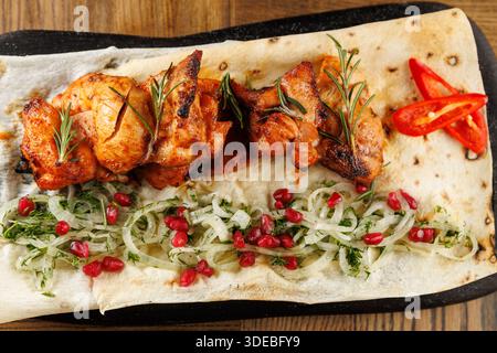 Saftige gegrillte Hähnchenspieße ruhen auf Fladenbrot, begleitet von einer Mischung aus frischen Kräutern, Zwiebeln und Granatapfelkernen auf einer Holzoberfläche. Stockfoto