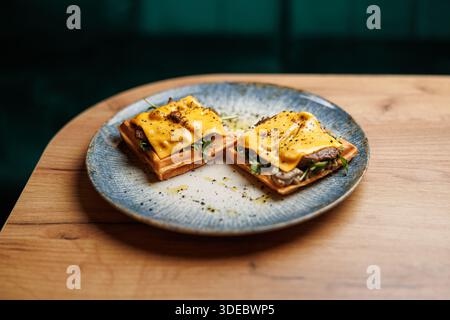 Zwei offene Sandwiches mit geschmolzenem Käse, Gemüse und Rindfleisch werden auf einer dekorativen blauen Platte platziert. Dieses Gericht wird auf einem Holztisch in einem serviert Stockfoto