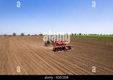 Die Vogelperspektive eines leuchtenden orangen Traktors bildet einen Kontrast zur braunen Erde und bereitet das Land für das Pflanzen unter einem riesigen blauen Himmel vor, Silistra, Bulga Stockfoto