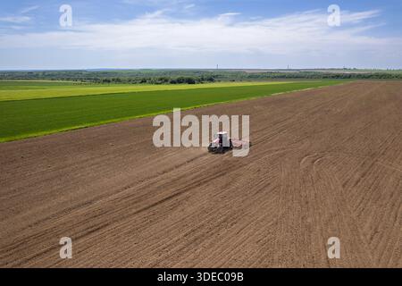 Aus der Vogelperspektive eines roten Traktors, der die Erde bebaut, im Kontrast zu den leuchtenden grünen Feldern unter einem weiten Himmel, Silistra, Bulgarien. Stockfoto