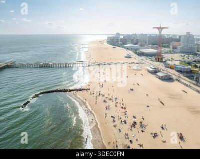 Aus der Vogelperspektive auf sonnenverwöhnten Sand treffen auf die rhythmischen Wellen des Ozeans, wobei der berühmte Fallschirmsprung die Skyline über der geschäftigen Coney Island Boa durchquert Stockfoto