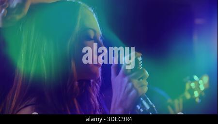 Singende Sängerin in dunklem Outfit mit Retro-Mikrofon auf der Bühne mit farbigen Lichtern, verschwommener Gitarre Stockfoto