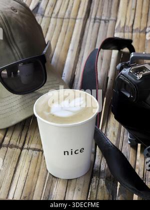 Blick auf den hohen Winkel heiße Latte Kaffeepapierbecher mit wunderschöner Milchschaumkunst in der Nähe von Digitalkamera Sonnenbrille Kappe auf Bambustisch während entspannender Ferien tra Stockfoto