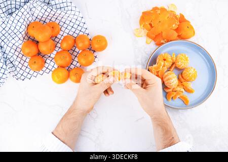 Draufsicht auf männliche Hände, die Mandarinensegmente auf einem weißen Marmortisch mit frischem Obst und blauem Teller trennen. Hochwertige Fotos Stockfoto
