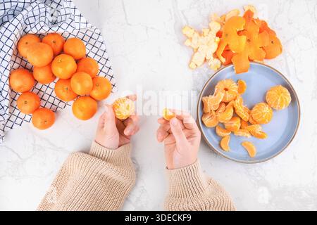 Blick von oben auf weibliche Hände, die geschälte Mandarinensegmente auf einem Marmortisch mit frischem Obst und blauem Teller halten. Hochwertige Fotos Stockfoto