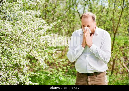 Allergie des Mannes mit saisonaler Allergie im Frühjahr im blühenden Garten im Frühling. Junger Mann, der niest und Nase bläst mit Nasentaschentuch vor blühendem Baum. Frühjahrsallergiekonzept Stockfoto