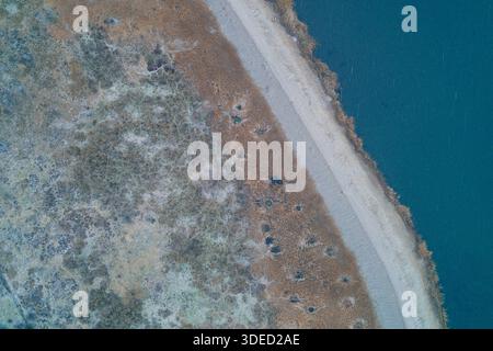 Von oben nach unten auf einen diagonalen Schotterdamm, der tiefblaues Wasser von einem verwelkten, frostigen Feuchtgebiet trennt. Eine strukturierte Landschaft, die eine Damm-Struktur, einen Staurand und Details des Wintersumpfs zeigt, die die Wasserwirtschaft darstellen. Stockfoto