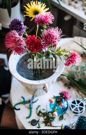 Violette und rosa Asterblüten stehen in Drahtgeflecht in der weißen Steinurne auf einem überdachten Arbeitstisch. Kreatives Wohlbefinden, Stressabbau durch Pflanzen Stockfoto