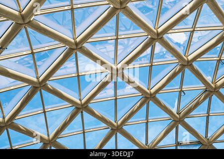 Geometrische Deckenstruktur aus Glas und Metall, die ein dreieckiges Gitter vor dem blauen Himmel bildet Stockfoto