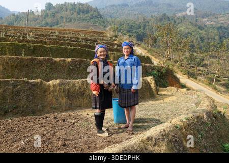 Lächelnde ethnische Frauen in traditioneller Kleidung auf Terrassenfeldern in ländlichen vietnamesischen Bergen, die lokale Kultur zeigen. Stockfoto