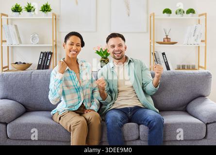 Fröhliches birassisches Paar, das auf dem Sofa sitzt, Sport im Fernsehen ansieht und den Sieg feiert Stockfoto