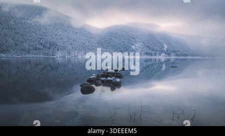 Wunderschöne Winter- und Seenlandschaft mit ruhiger Reflexion auf dem Kootenay Lake mit Sonnenaufgang oder Sonnenuntergang Licht und Nebel in Nelson, British Columbia, CA Stockfoto