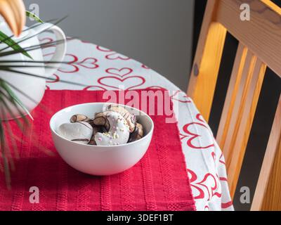 Weiße Schüssel mit verschiedenen dekorierten Keksen auf einem roten Tischset und einer Tischdecke mit Herzmuster. Mischung aus sichelförmigen, herzförmigen, glatten und schokoladenüberzogenen Keksen Stockfoto