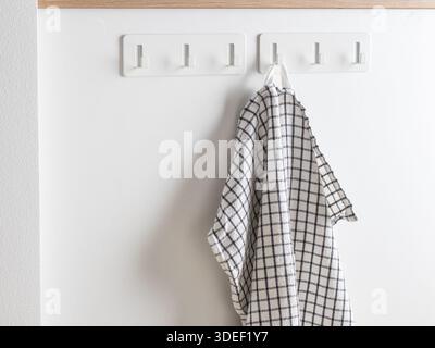 Schwarz-weiß kariertes Küchentuch aus Baumwolle, das an einem weißen selbstklebenden Wandhaken hängt. Saugfähiges, langlebiges Geschirrtuch im Bauernhausstil zum Trocknen von Geschirr Stockfoto