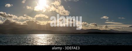 Panoramablick über die Straße von Menai in Richtung der Berge von Carneddau, mit niedrigen Wolken und Sonnenstrahlen, die durch einen dramatischen Himmel brechen Stockfoto