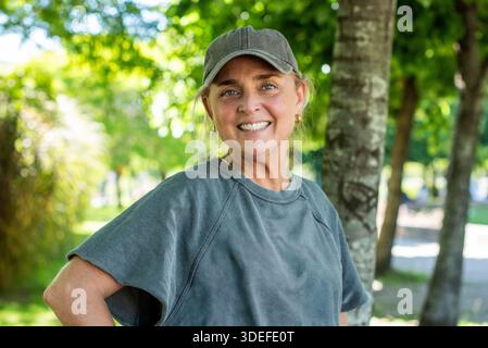 Lächelnde Frau mittleren Alters mit Mütze und Sportbekleidung in einem sonnigen Park Stockfoto