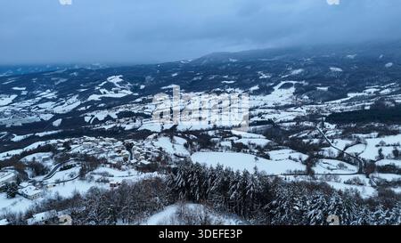 Eine atemberaubende Luftperspektive fängt ein ruhiges italienisches Dorf mit unberührtem Schnee ein, umgeben von sanften Hügeln und fernen Bergen Stockfoto