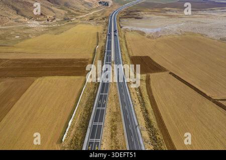 Aus der Vogelperspektive einer geraden Autobahn durchzieht die trockene Landschaft und schafft einen starken Kontrast zwischen dem dunklen Asphalt und dem umliegenden goldenen Feld Stockfoto