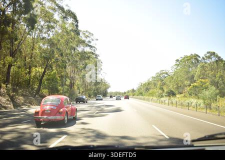 Roter Volkswagen Käfer fährt mit anderen Autos auf einer Autobahn, umgeben von Bäumen im Hintergrund. Stockfoto