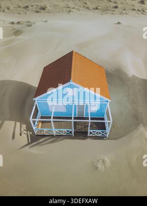 Blick aus der Vogelperspektive auf ein einsames blau-weißes Strandhaus mit einem lebhaften orangen Dach, eingebettet in die hügeligen Sanddünen, Costa da Caparica, Setubal, Stockfoto