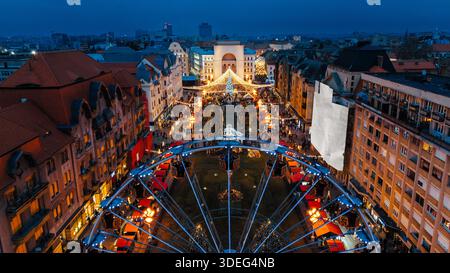 Aus der Vogelperspektive auf den lebhaften Weihnachtsmarkt, ein Riesenrad im Vordergrund und den beleuchteten Siegesplatz, Timișoara, Timiș, Rumänien. Stockfoto