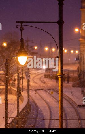 Budapest, Ungarn - 6. Januar 2026: Eine leuchtende Vintage-Straßenlaterne steht im Vordergrund, während Schnee über eine verschwommene Budapester Straßenbahnlinie fällt Stockfoto