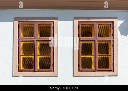 Zwei Fenster im Vintage-Stil mit Holzrahmen und strukturierten bernsteinfarbenen Glasscheiben, die in vier Abschnitte unterteilt sind, auf einer weißen Fassade mit braunem Rahmen. Stockfoto