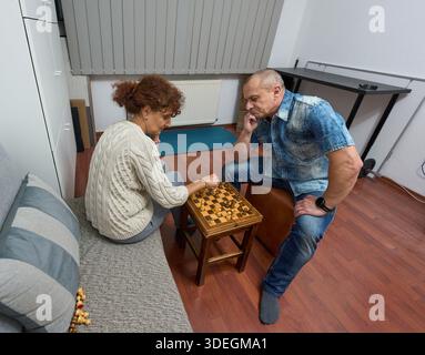 Paare mittleren Alters spielen gemeinsam auf Holzbrett im Wohnzimmer zu Hause Schach Stockfoto