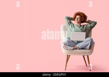 Rothaarige Freiberuflerin mit Laptop im Sessel auf rosa Hintergrund Stockfoto